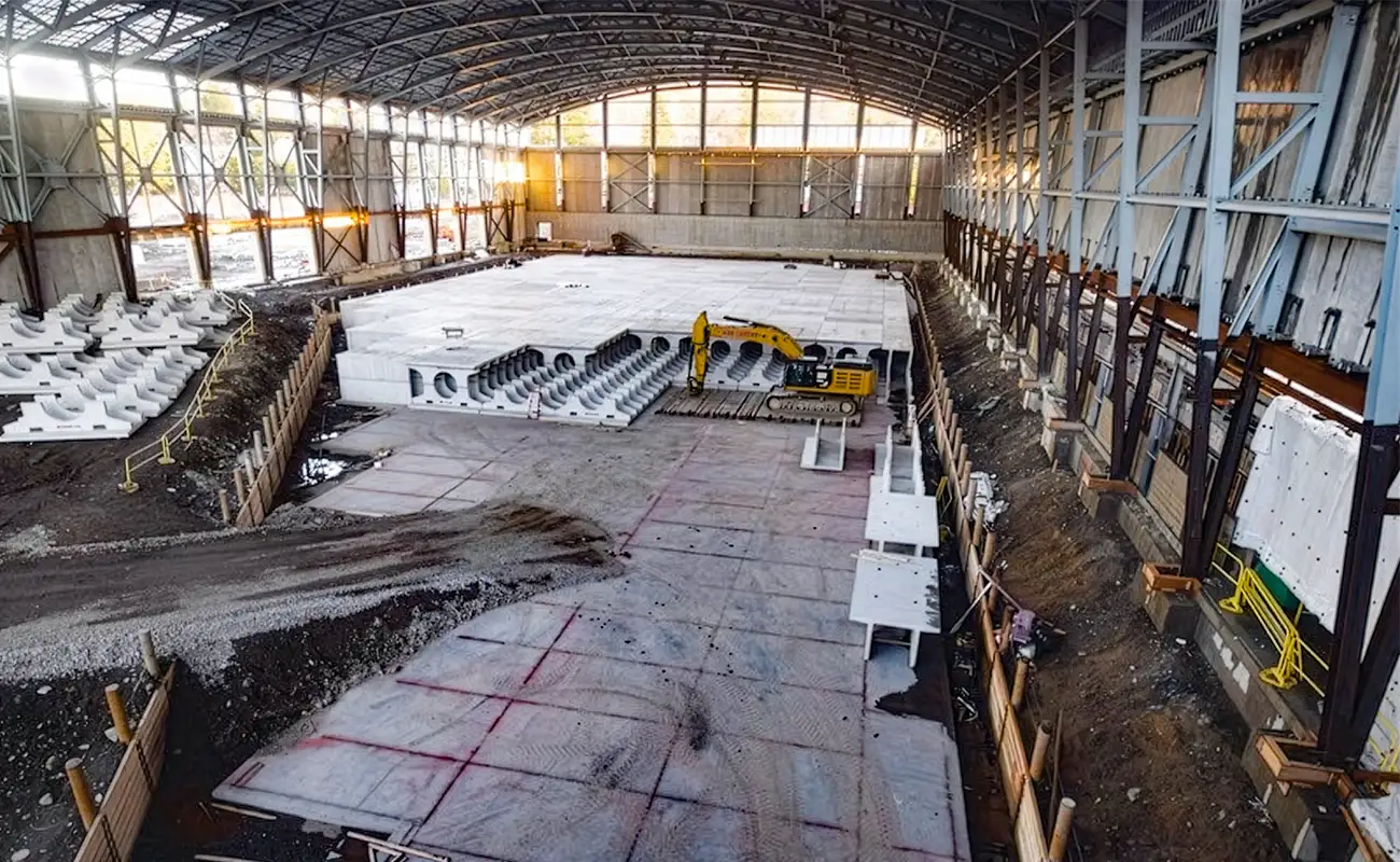 A large indoor construction site with a partially completed concrete floor, structural steel beams, and a yellow excavator working near precast concrete segments.