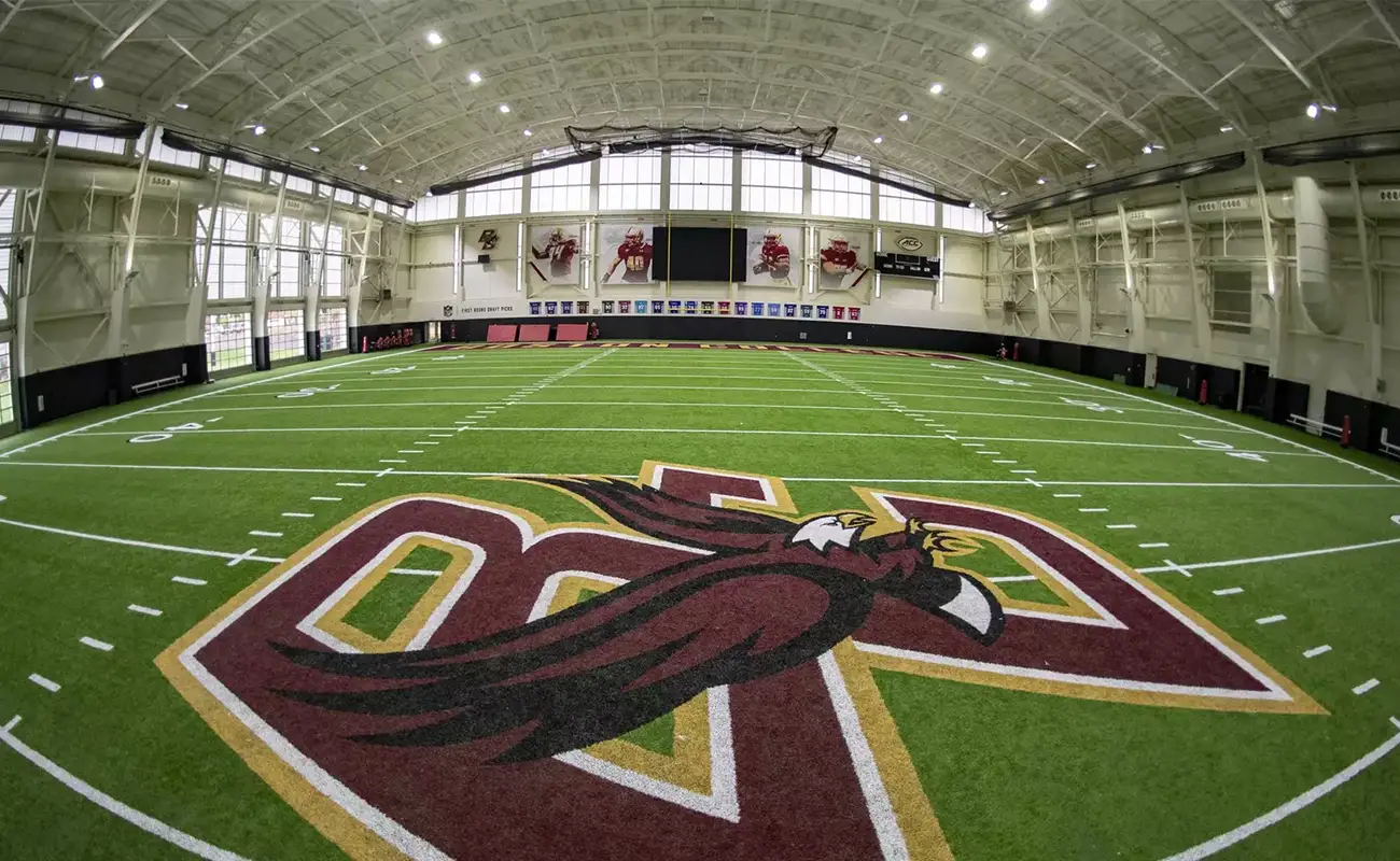Indoor football field with artificial turf, large maroon and gold eagle logo at midfield, and sports banners hanging on the back wall.
