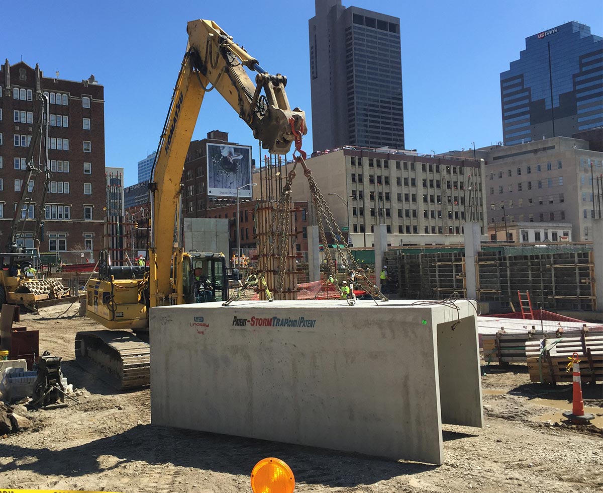 Construction site with heavy machinery moving large concrete slabs. Workers are visible, and city buildings are in the background under a clear blue sky.