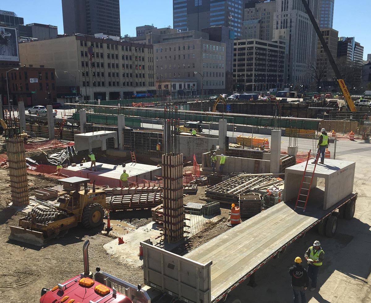 Construction site with workers, machinery, and trucks. Steel and concrete materials are visible. Buildings and a crane are in the background.