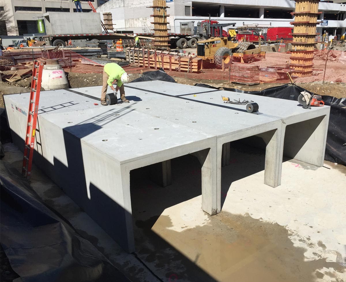 Construction worker on a large concrete slab with open compartments, surrounded by construction materials, ladders, and equipment on a worksite.