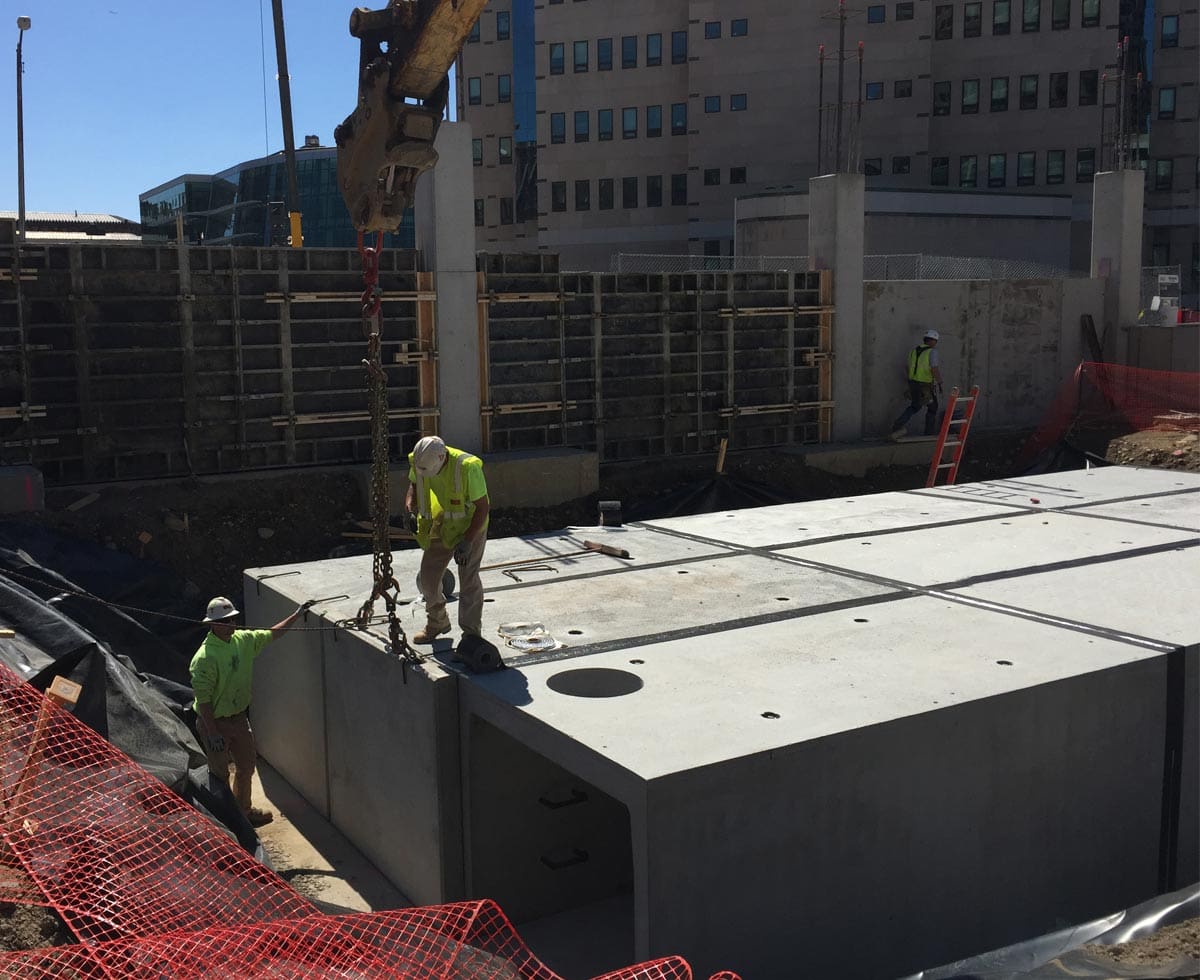 Construction site with workers guiding a large concrete block into place using a crane. Plaid and mesh barriers surround the area, with a tall building in the background.