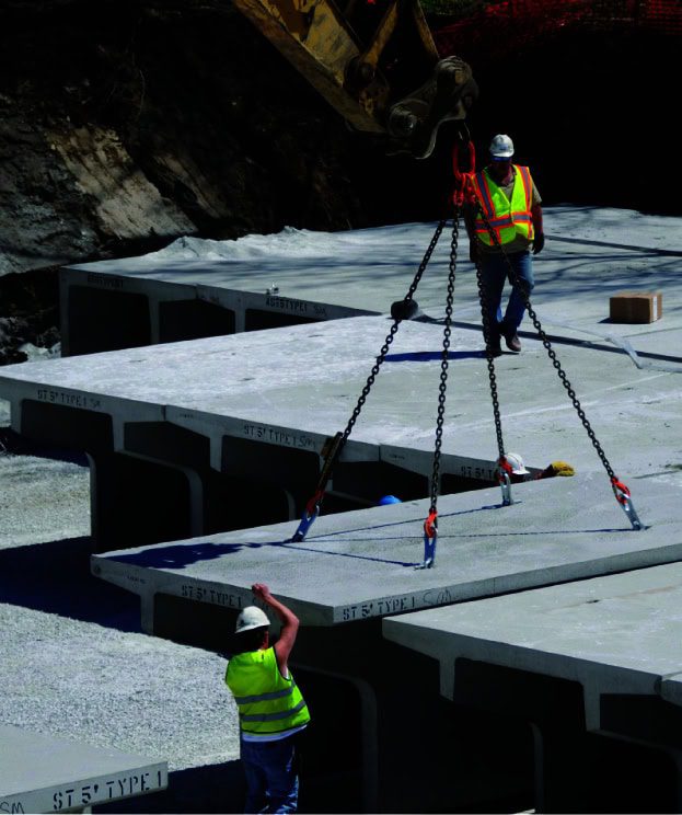 Construction workers in safety gear expertly maneuver large concrete slabs with a crane at a bustling construction site.