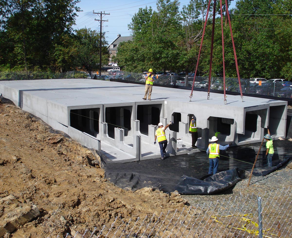 Construction workers guide the installation of large concrete structures at a site surrounded by trees and a fence.