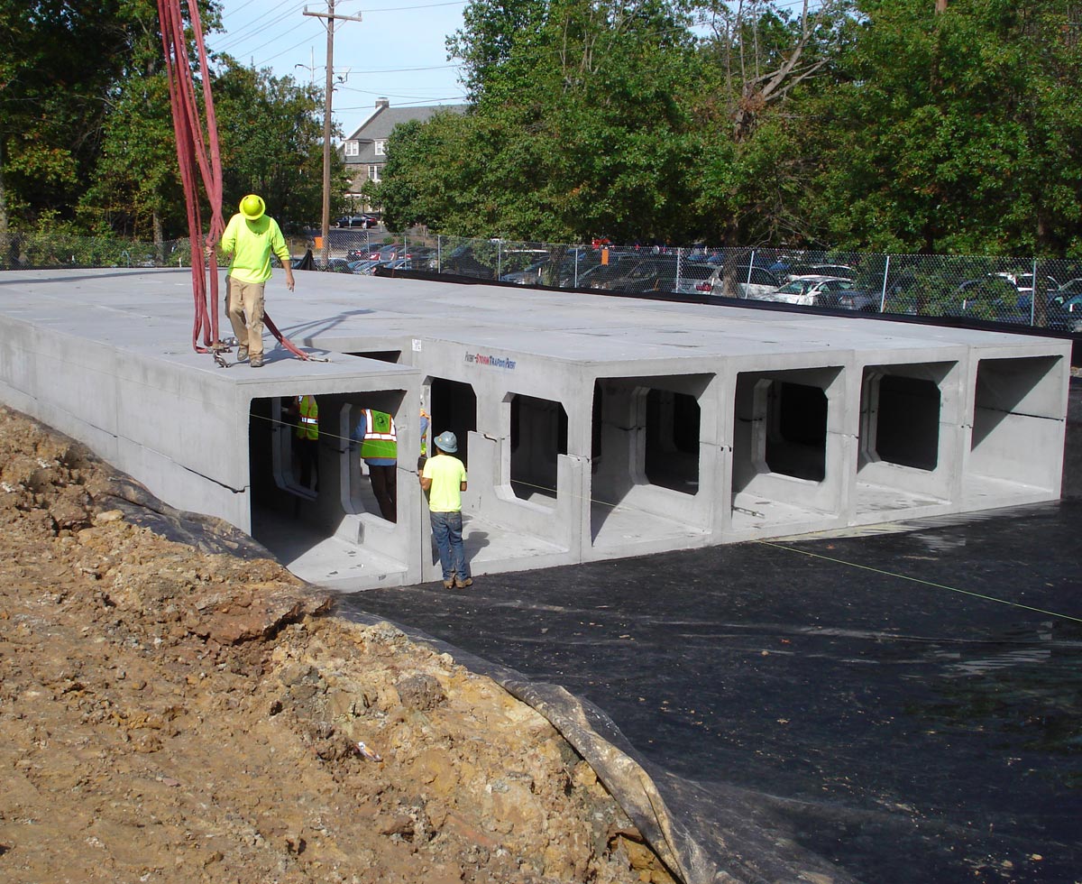 Construction workers assembling large concrete box culverts at a construction site surrounded by trees and power lines.