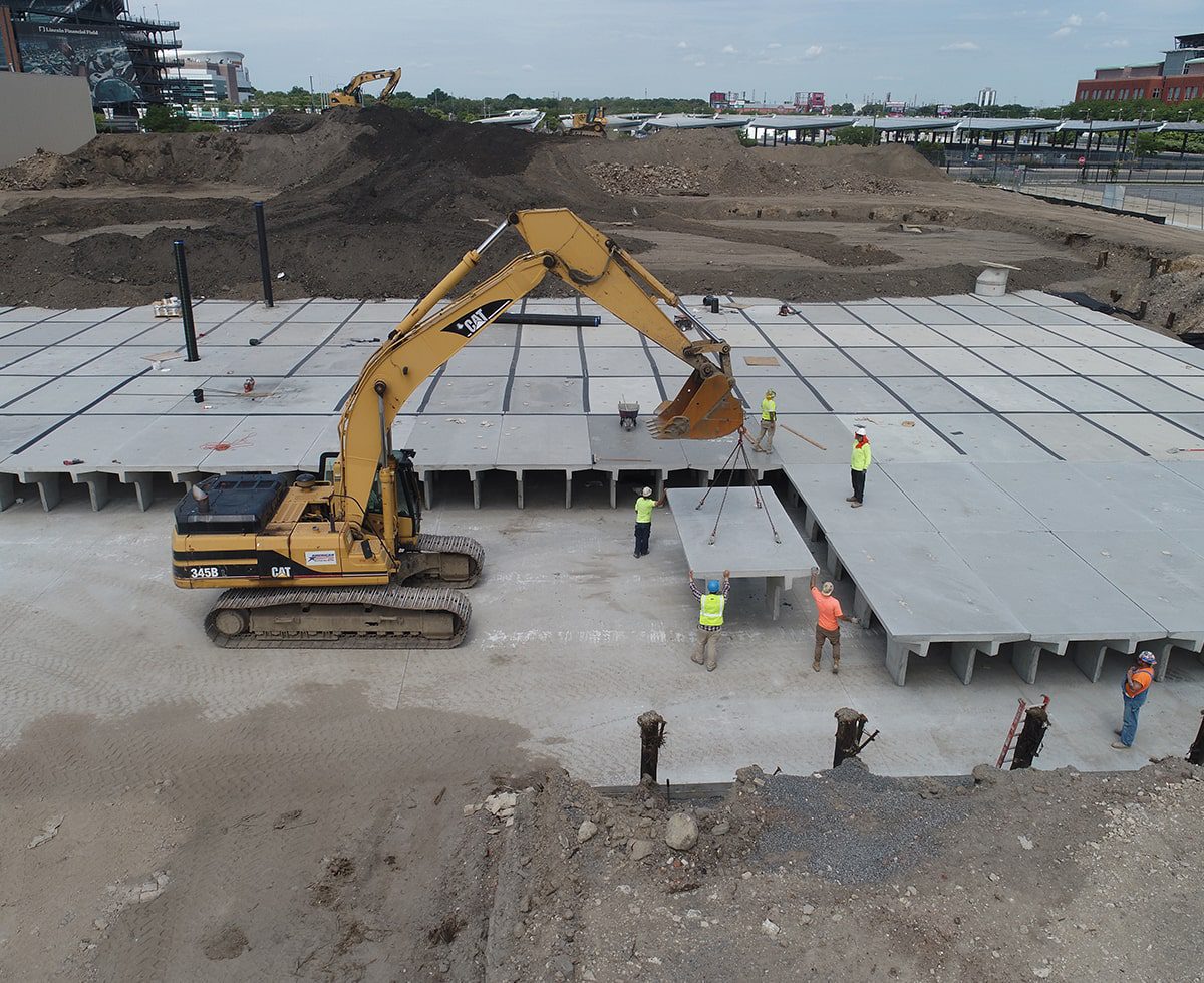 An excavator methodically places massive concrete slabs at a bustling construction site, guided by diligent workers clad in bright safety gear.