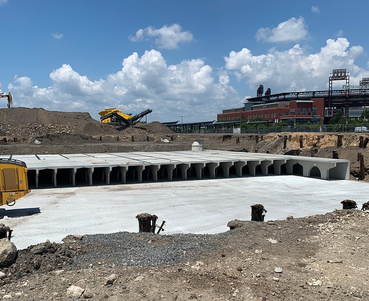 A construction site features large concrete culverts scattered across a cleared area. Heavy machinery looms beside mounds of dirt in the background, while a lone building stands under a partly cloudy sky.