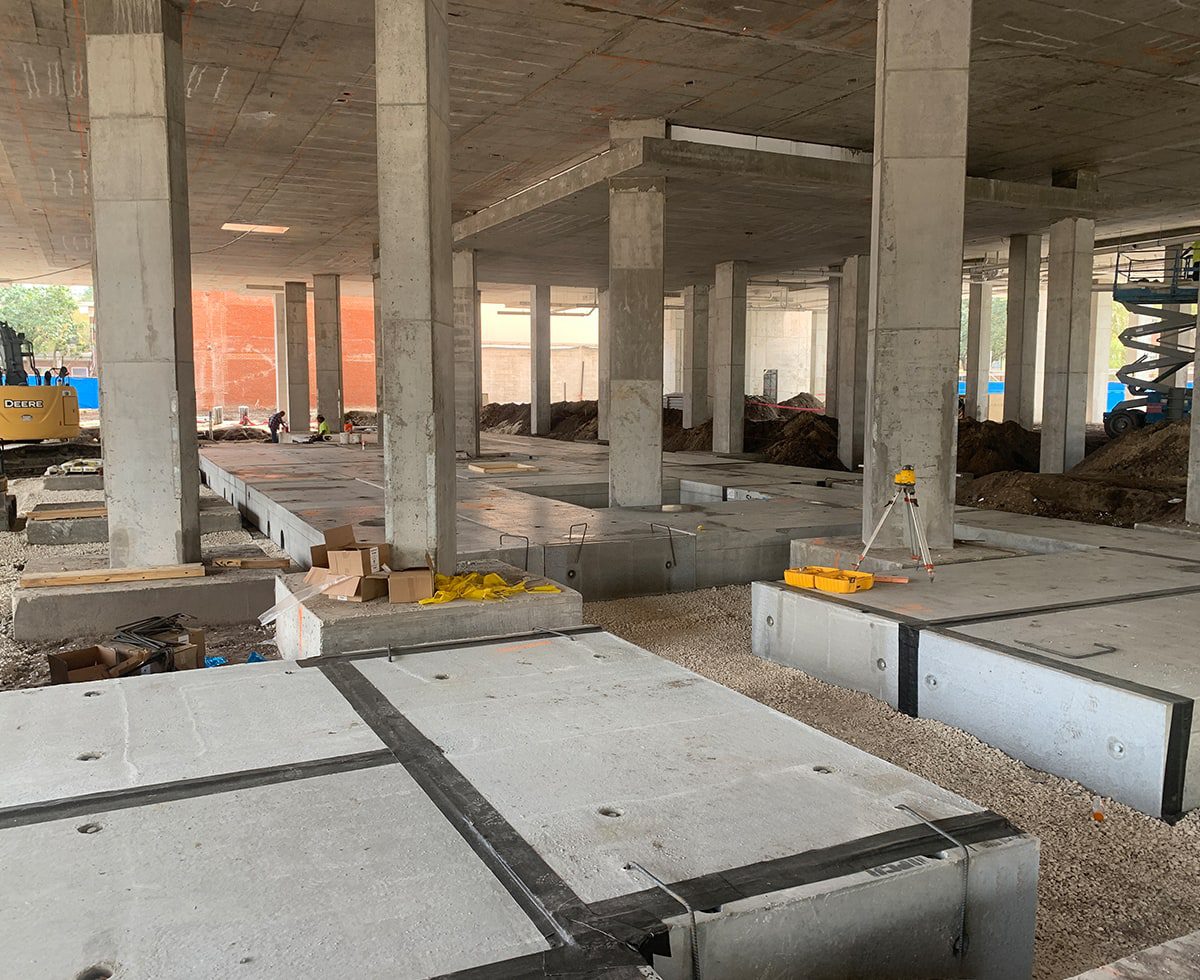 Construction site with concrete slabs and support pillars under a building's structure. Equipment and materials are scattered around.