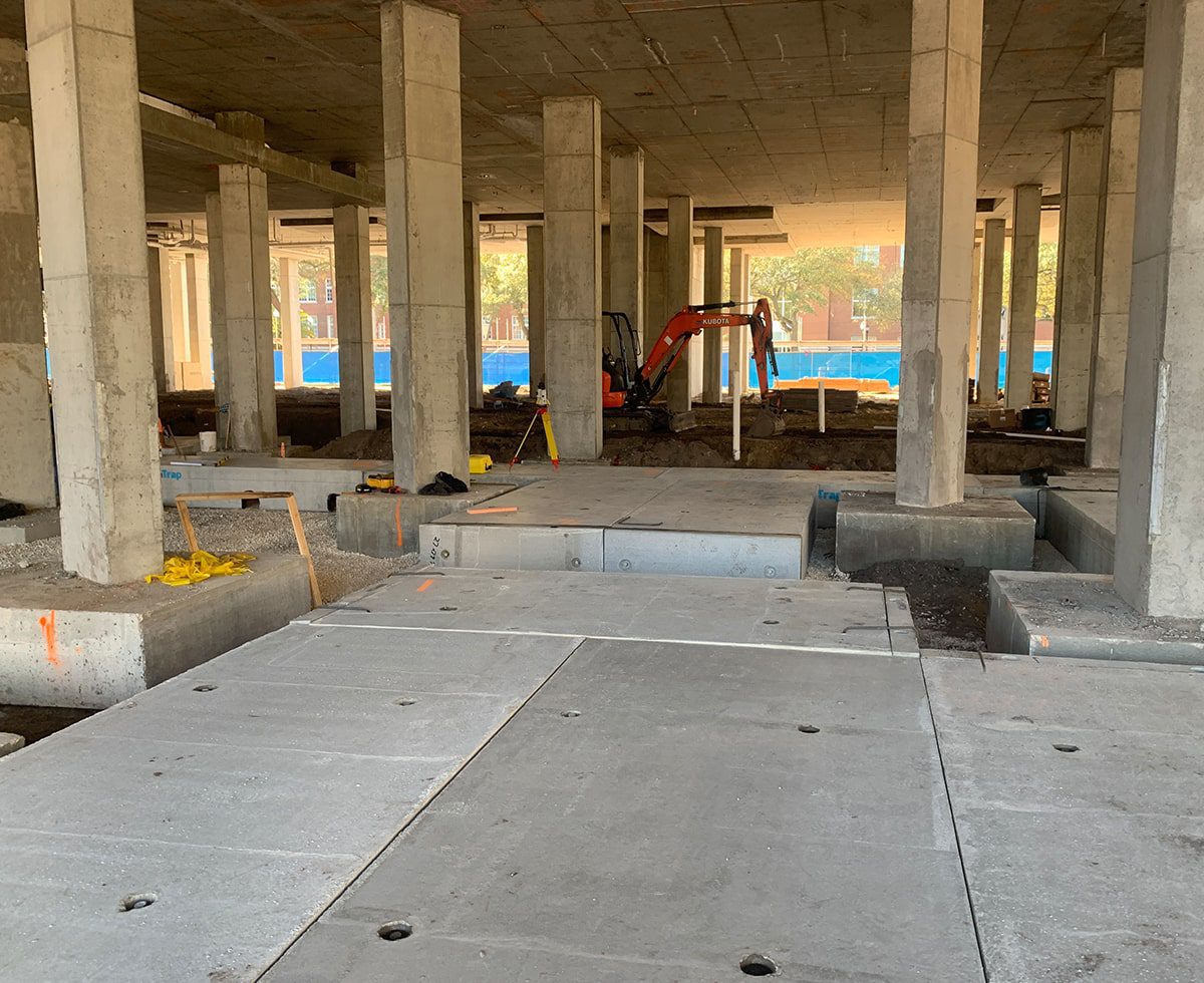 Construction site under a building with concrete slabs, columns, and a small excavator in the background.
