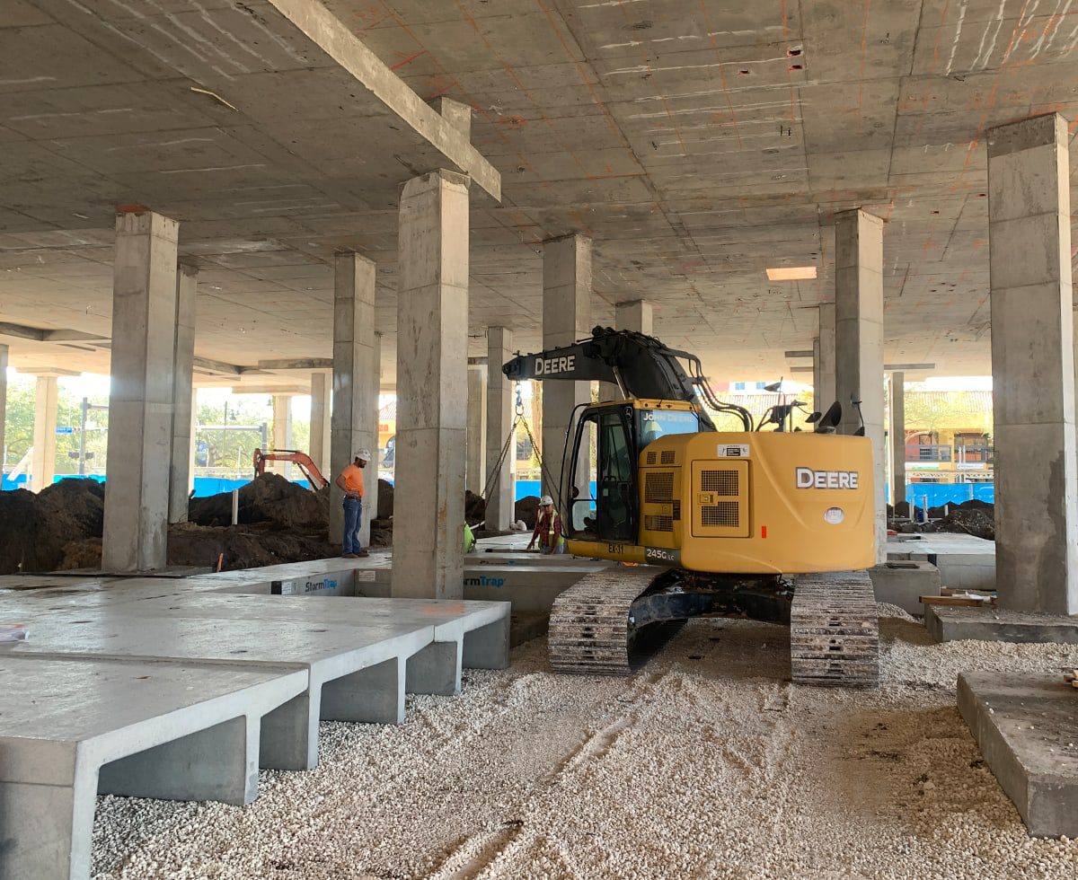 A bustling construction site features a yellow excavator under a partially completed building. Workers in hard hats are diligently moving around, surrounded by concrete pillars and gravel scattered across the ground.