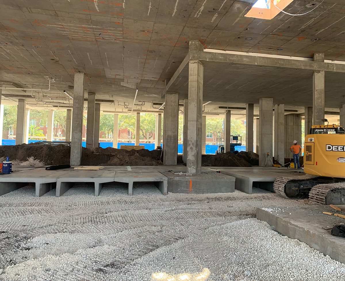 Construction site with exposed concrete columns and beams, piles of dirt, and a yellow excavator. Construction workers in hard hats are present. Blue fencing is visible outside.