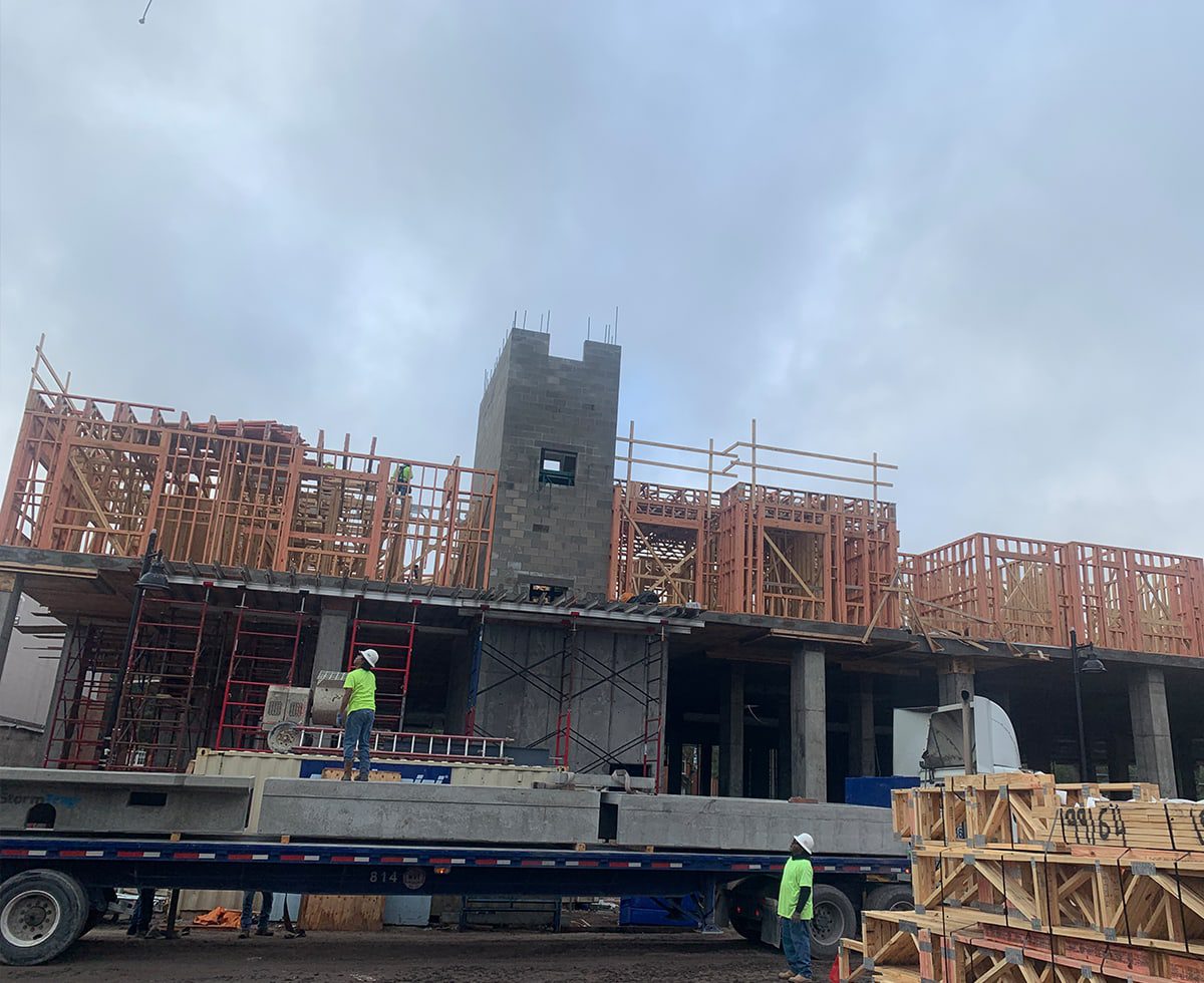 Construction site with workers building a multi-story structure using wood and cement. A flatbed truck is parked in front, and overcast skies are visible above.