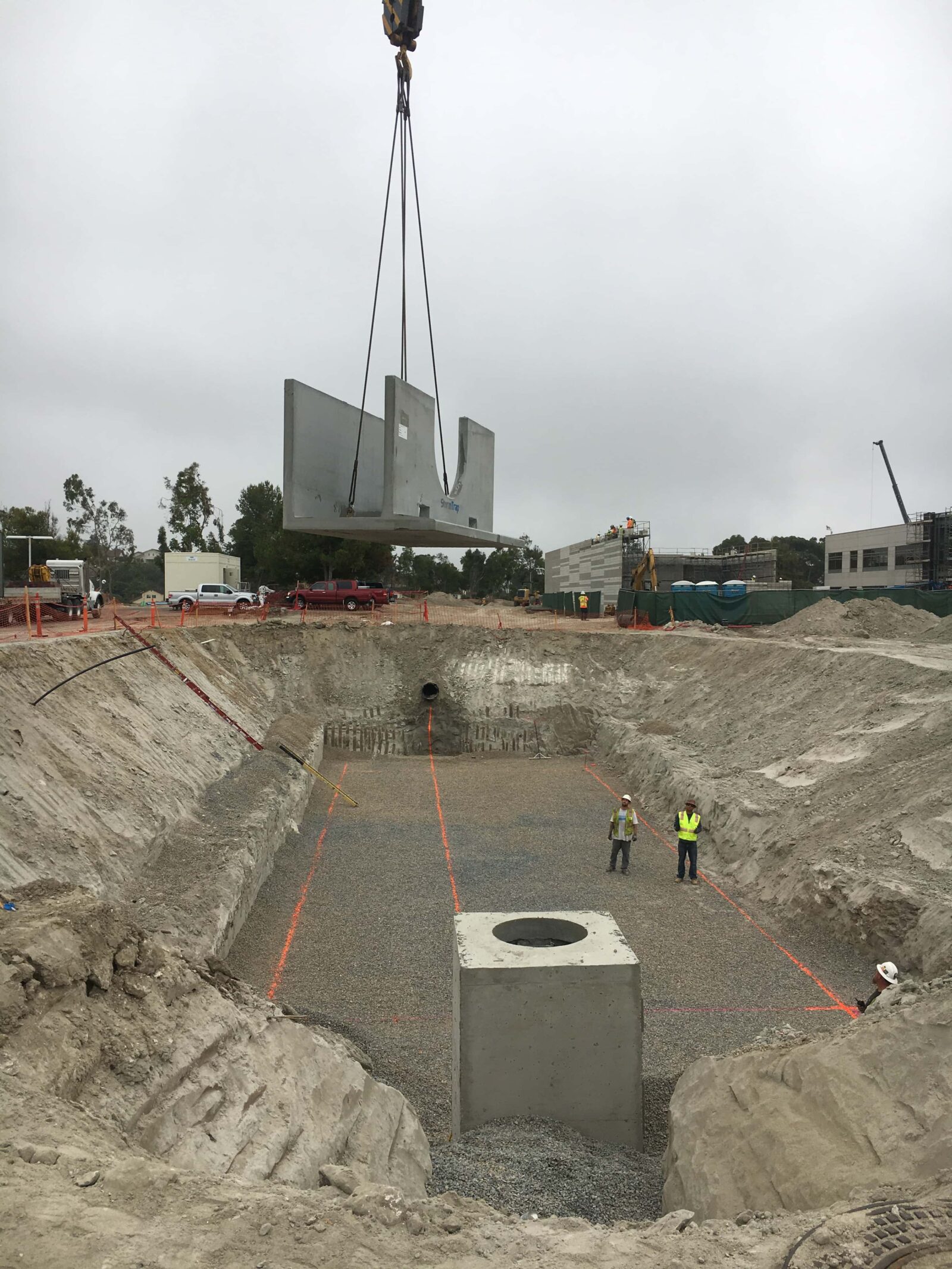 Construction site with a large concrete panel being lowered by a crane into a pit. Two workers in hi-vis vests stand nearby, supervising the operation.