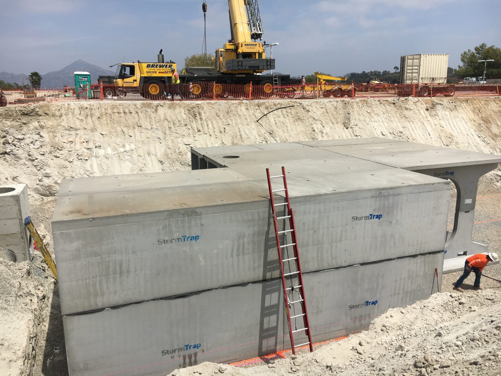 Construction site with large concrete stormwater tanks being installed. A crane is positioned nearby, and a worker in an orange vest is visible. A ladder leans against the tanks.