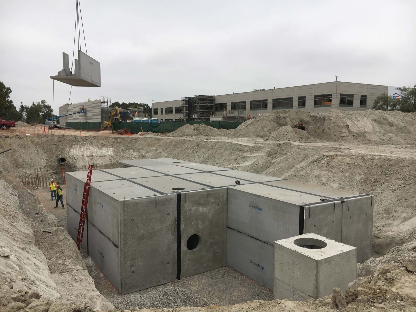 Construction site with a large, partially installed concrete water detention system. A crane lifts a concrete piece. Workers and building in the background.