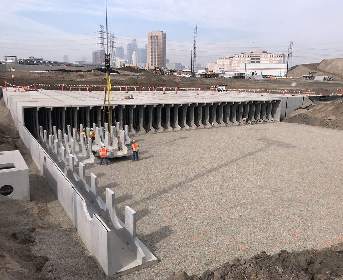 Construction site with workers assembling large concrete structures on a gravel surface, with city skyline and machinery in the background.