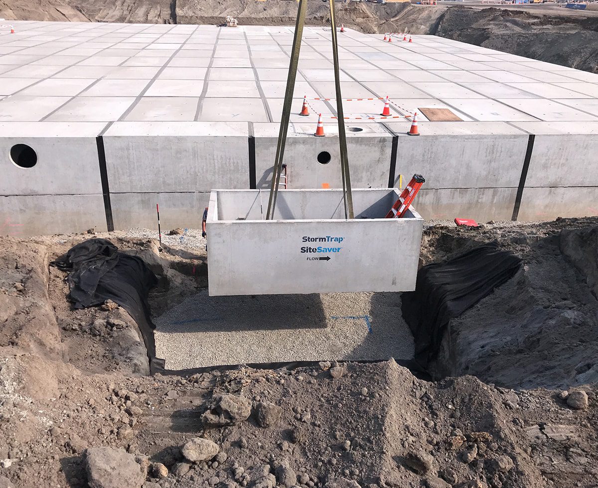 A concrete structure labeled "StormTrap SiteSaver" is being lowered into a trench at a construction site. The area is surrounded by dirt and construction cones are visible on the ground.