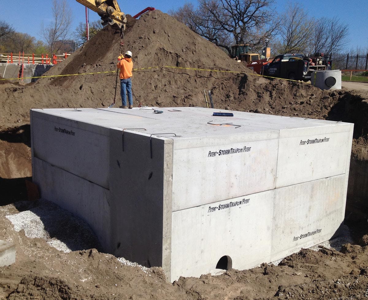 A worker stands near a large concrete stormwater vault being installed at a construction site, with an excavator in the background. Gravel is spread below the vault, and earth surrounds it.