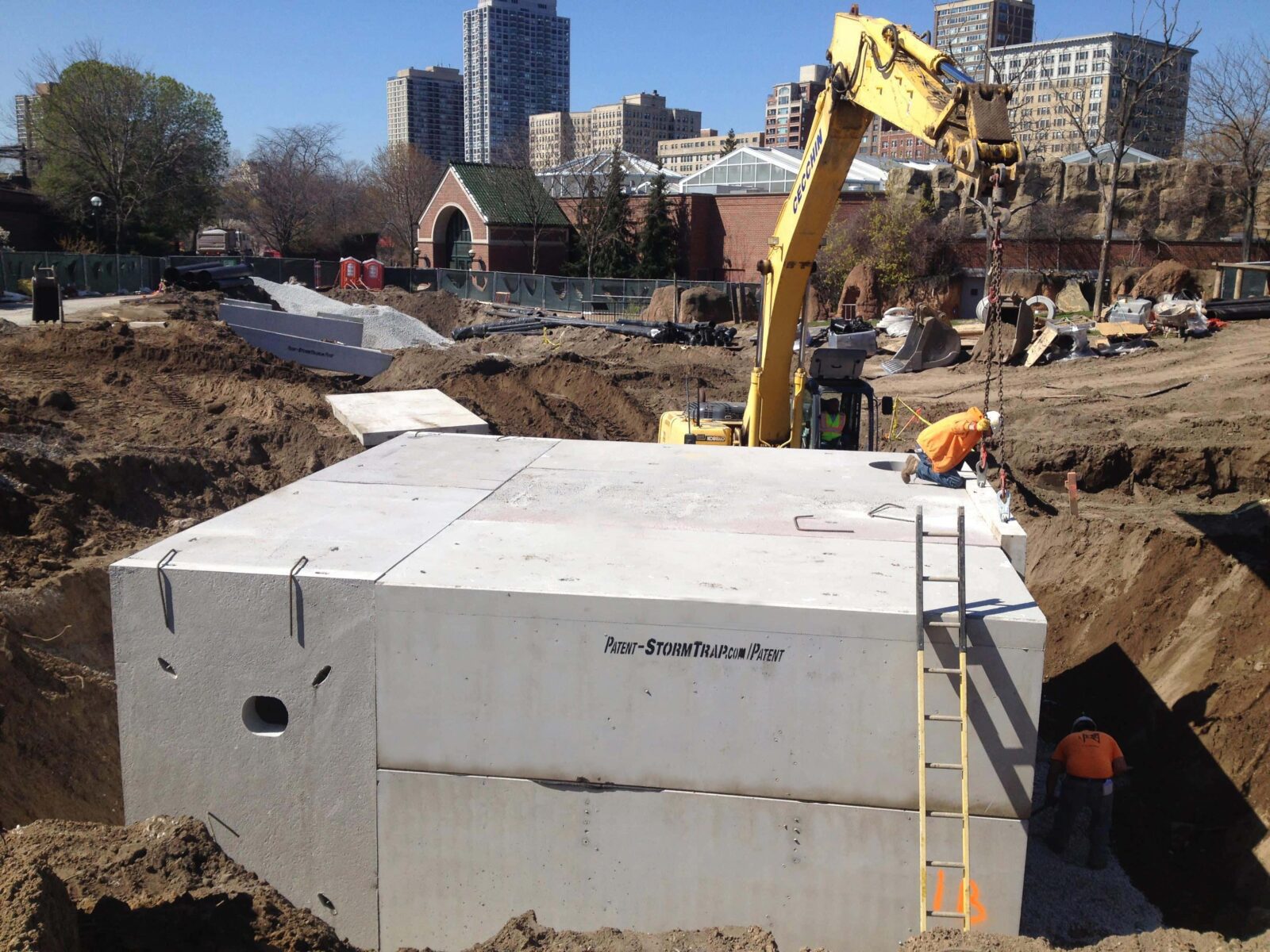A construction site with workers installing a large concrete stormwater tank using an excavator. Nearby are buildings and trees.