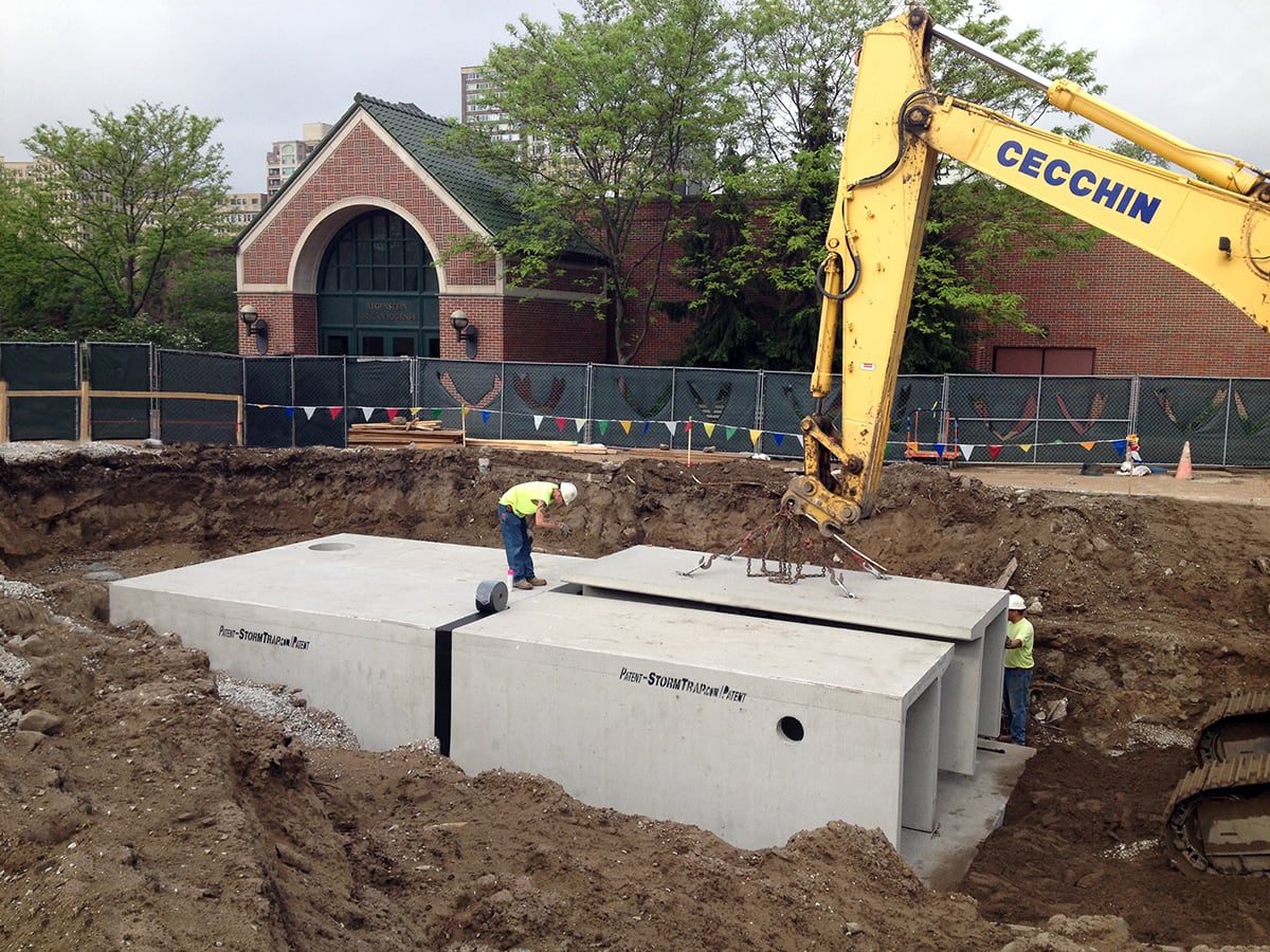 Construction site with workers installing large concrete structures using an excavator. A brick building and trees are visible in the background.