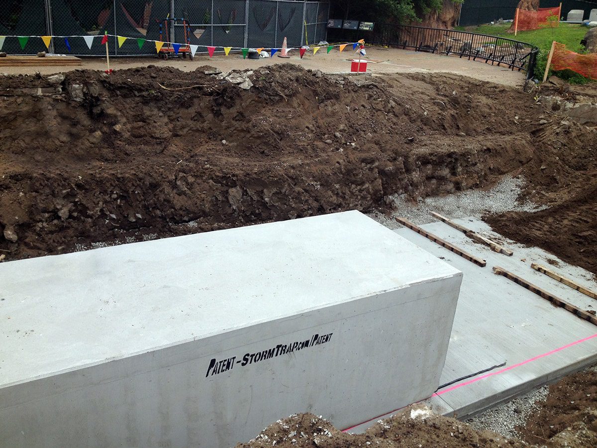 Construction site with a large concrete storm trap in a trench, surrounded by soil and construction barriers.