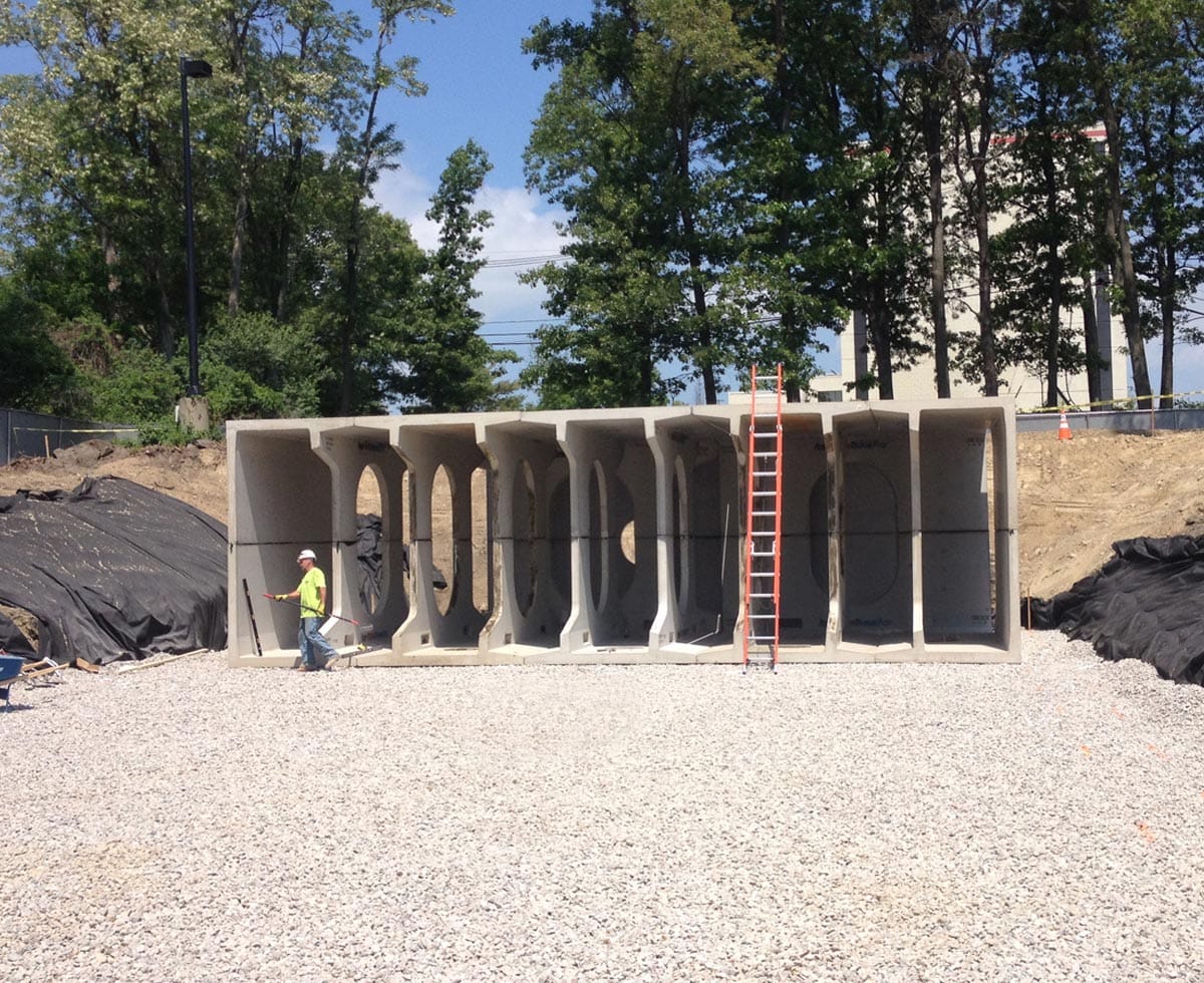 Large concrete box culverts on a gravel site with a ladder leaning against them; a worker in a neon vest stands nearby; trees in the background.