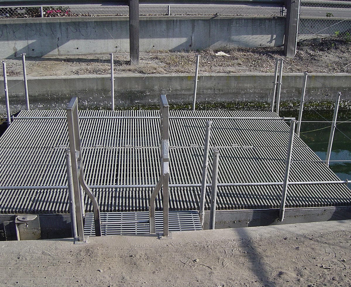A metal grate structure with a fence and railing is positioned over a water channel beside a dirt path and a road.