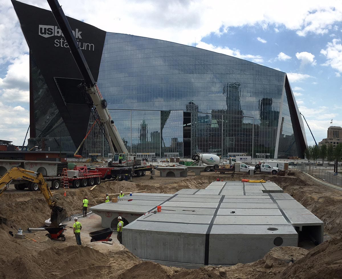 Construction site with workers and machinery near a large reflective glass building marked "U.S. Bank Stadium.