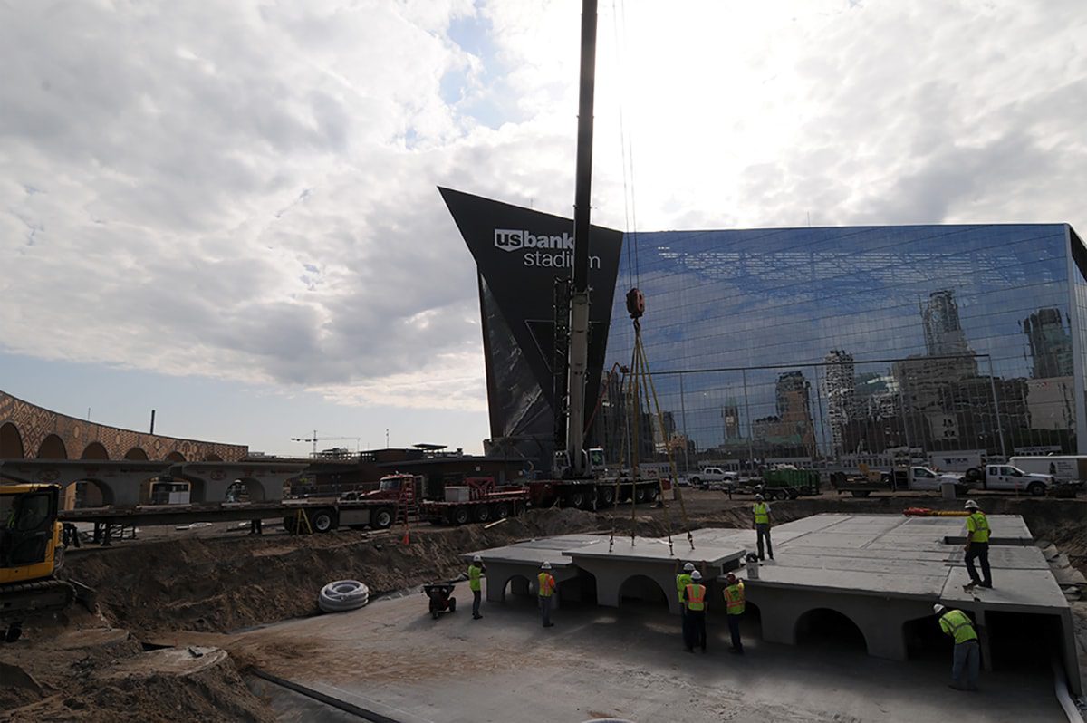 Construction workers and machinery at a site near a glass building labeled "US Bank Stadium" under a partly cloudy sky.