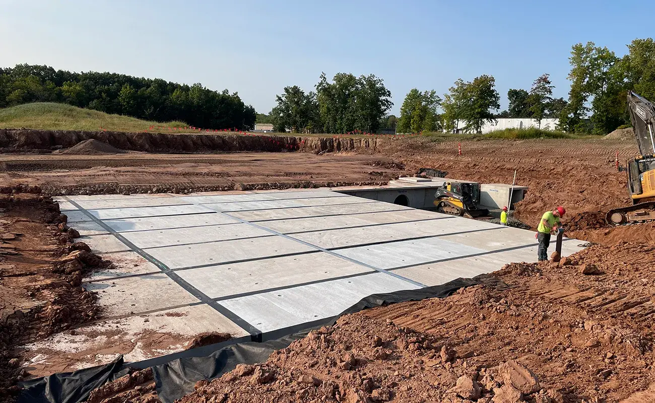 Two construction workers stand near large, rectangular concrete slabs being installed in a dug-out area at a construction site, with trees and machinery in the background.