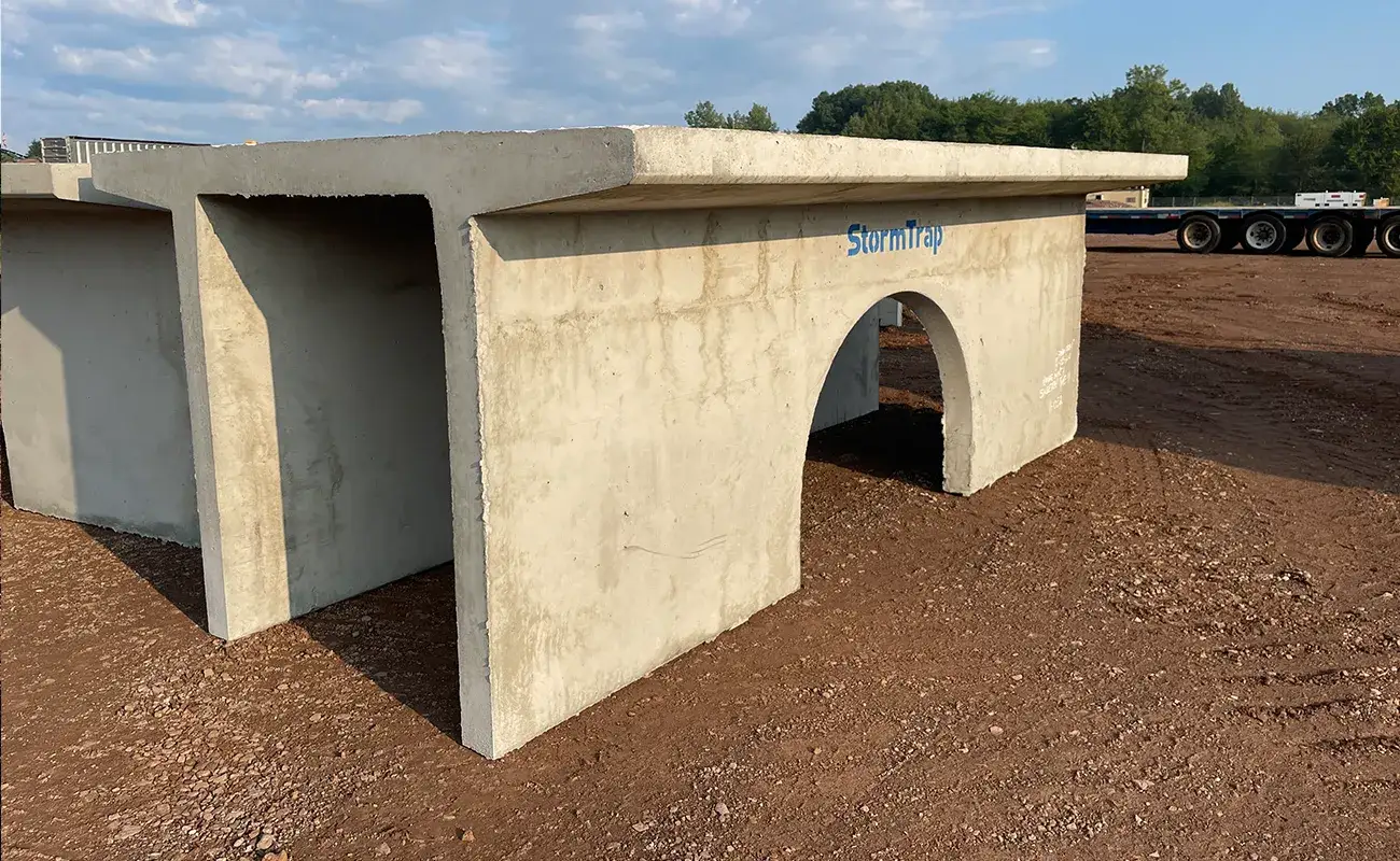 A large precast concrete box culvert sits on a dirt lot, featuring a rectangular shape with an arched opening and the word "StormTrap" on its side.