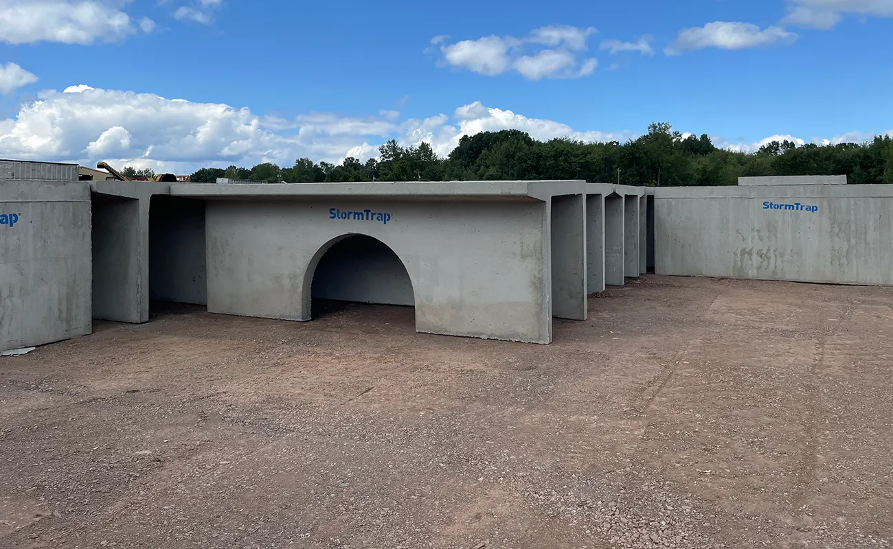 Large precast concrete stormwater management structures labeled "StormTrap" are stacked outdoors on a dirt lot under a partly cloudy sky.