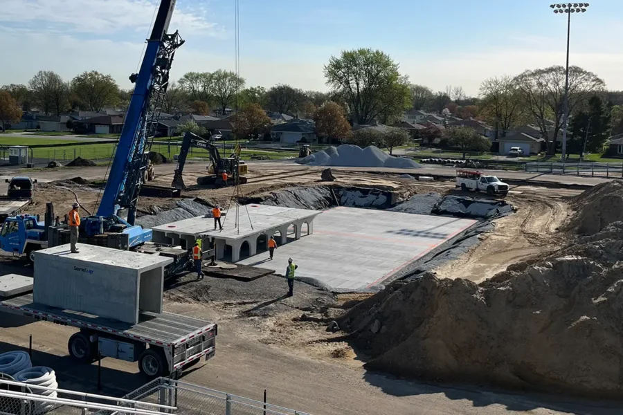 Construction workers and heavy machinery installing large concrete structures at an outdoor site with dirt piles, trucks, and a field in the background.