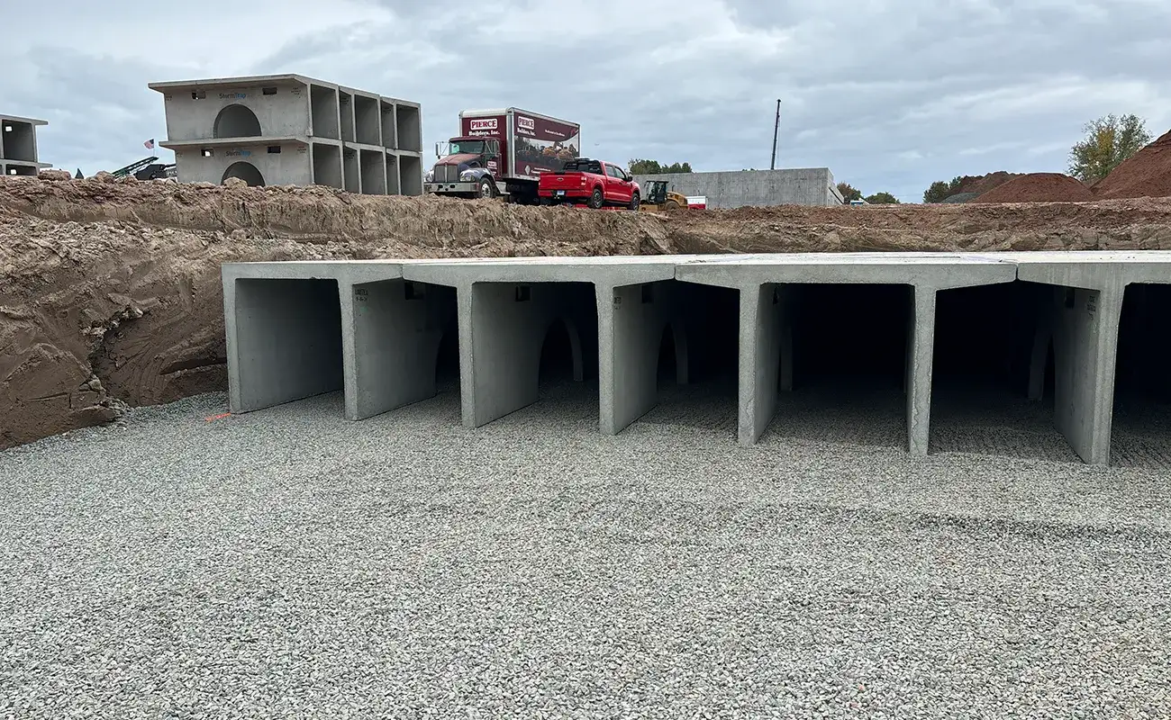 Large concrete box culverts are installed in a gravel bed at a construction site, with trucks and additional concrete structures in the background.