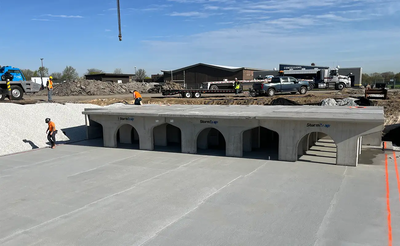 Construction site with workers installing large precast concrete stormwater vault sections into an excavated area, surrounded by gravel and machinery under a clear sky.