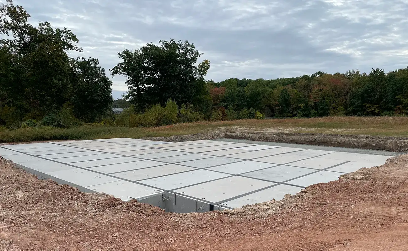 A foundation slab made of precast concrete panels is installed in a cleared area surrounded by dirt and trees under a cloudy sky.