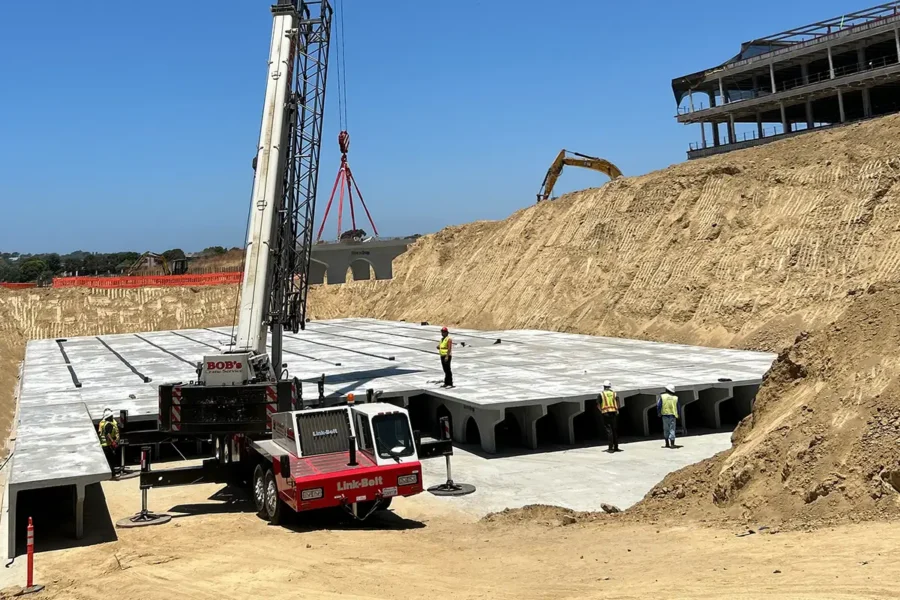 Construction site with workers, a crane, and large concrete structures being installed in an excavated area; unfinished building in the background.
