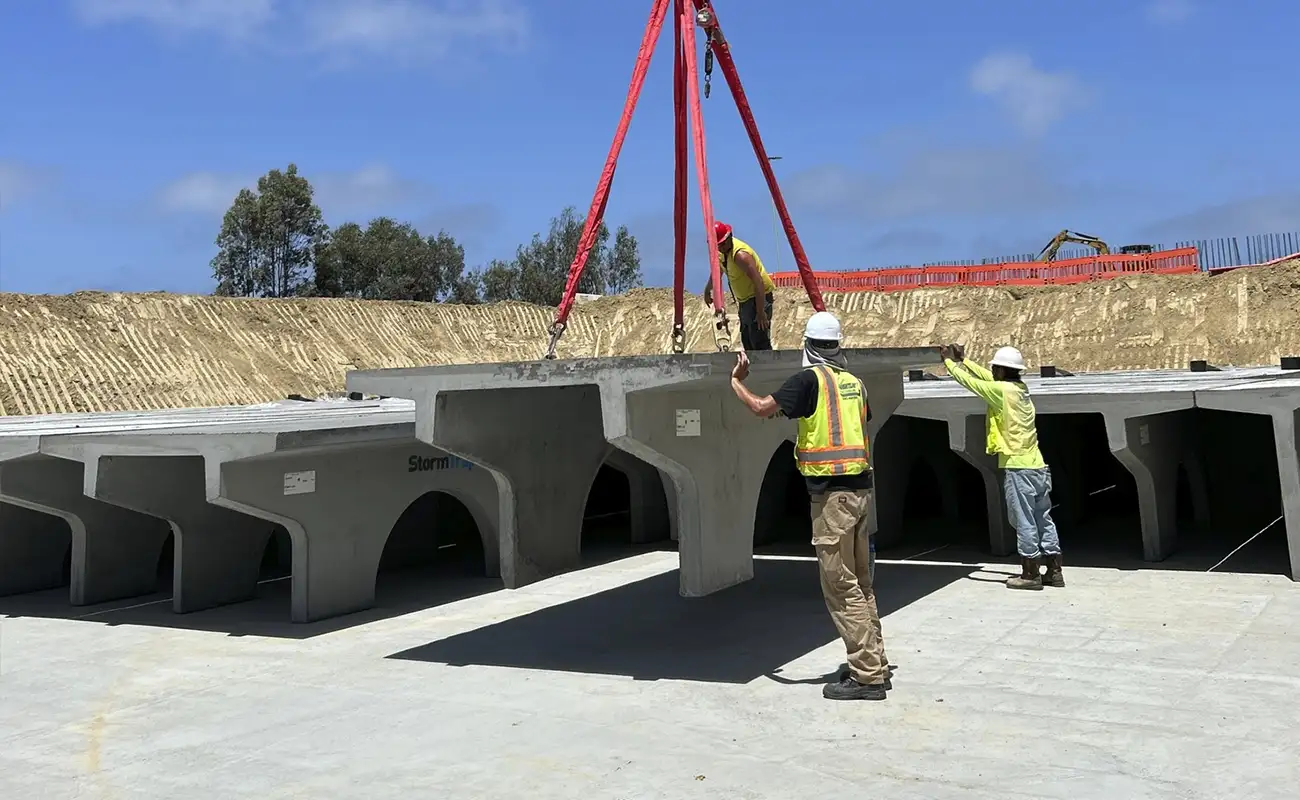 Construction workers guide a crane as it lifts and positions a large concrete stormwater structure at a construction site under a blue sky.