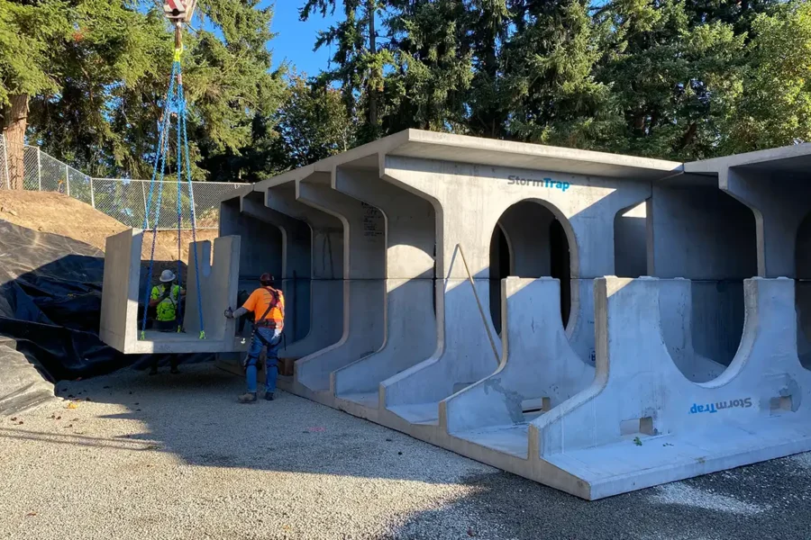 A construction worker guides a large precast concrete stormwater management chamber being lifted by a crane at an outdoor work site.