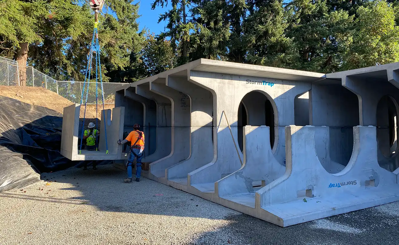 A construction worker guides a large precast concrete stormwater management chamber being lifted by a crane at an outdoor work site.