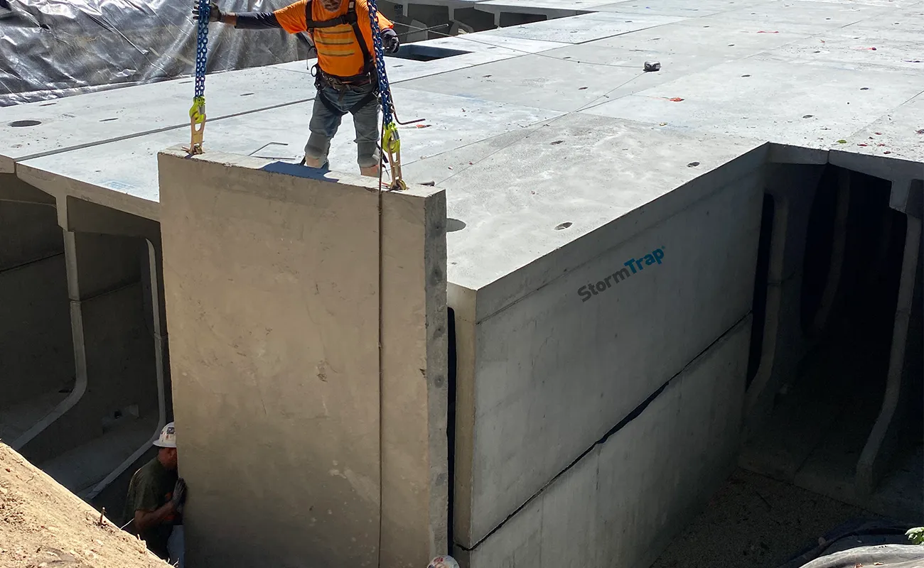 A construction worker guides a large concrete panel into place at an industrial site, with another worker partially visible below.