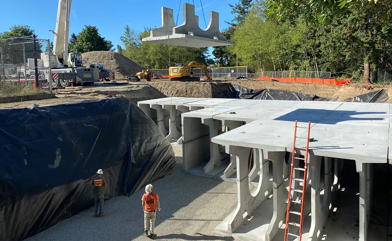 A crane lowers a large concrete slab onto an underground structure at a construction site, with workers in safety gear observing the process.