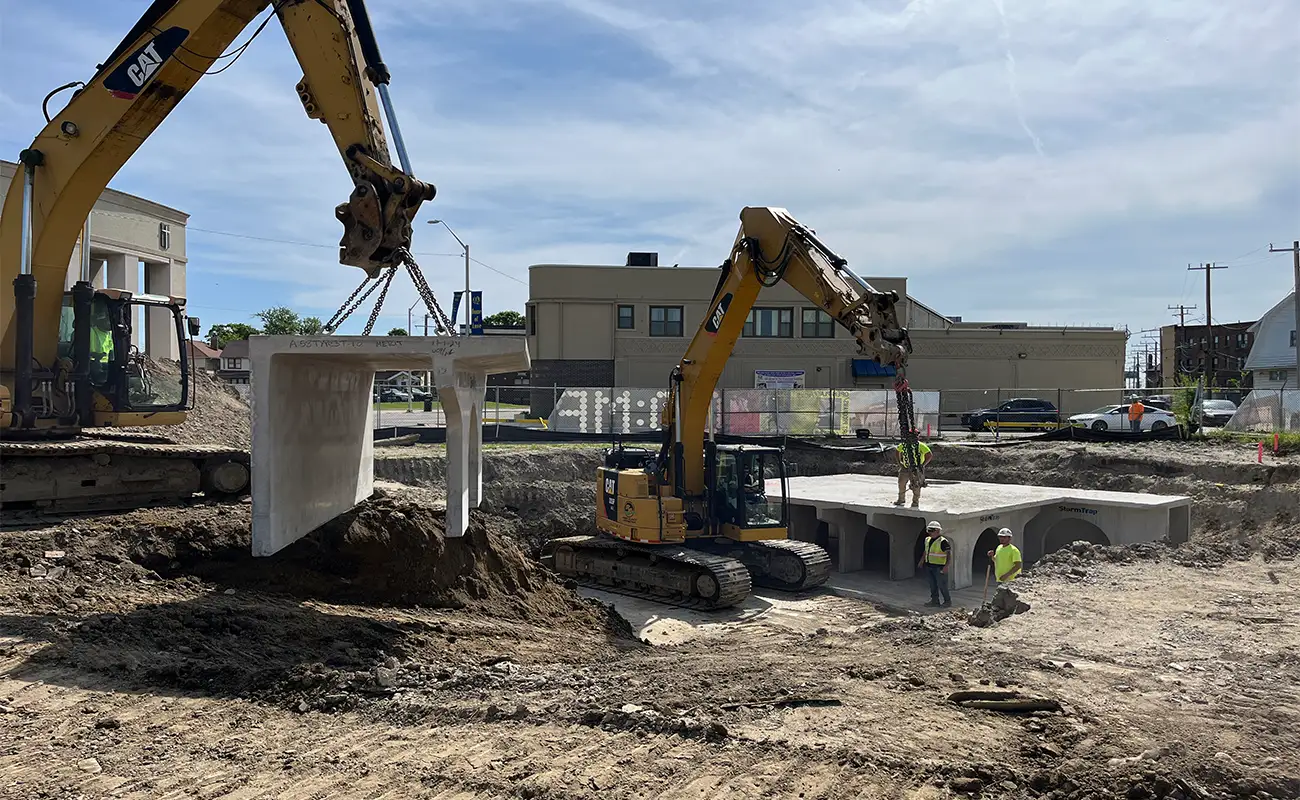 Two excavators lift and position large concrete structures while construction workers guide them at an urban construction site.