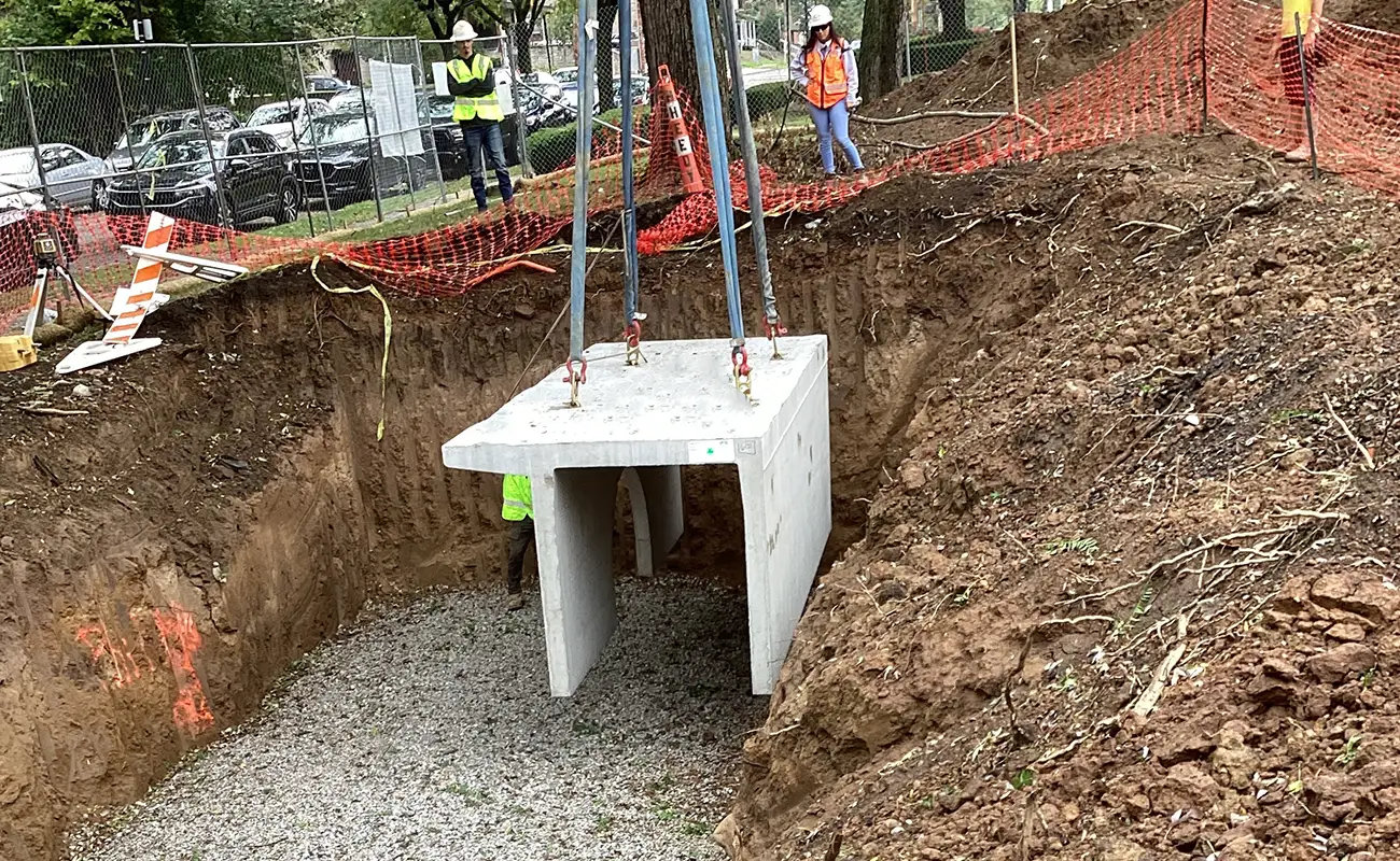 A large concrete drainage structure is being lowered into a dug trench by a crane at a construction site, with workers in safety gear observing nearby.