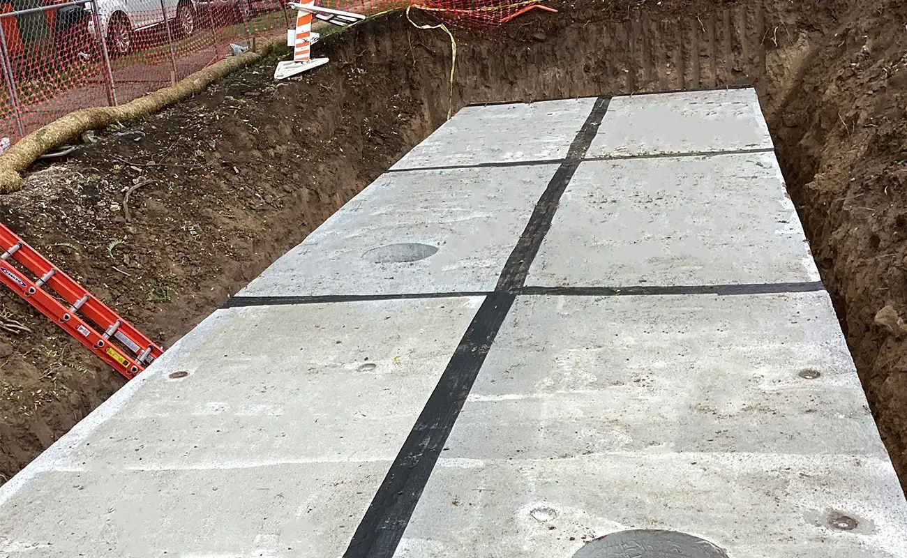 Large concrete slabs with visible seams are installed in a rectangular excavation site, surrounded by dirt, construction barriers, and a red ladder.