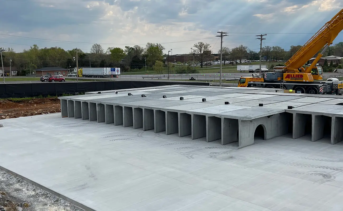 Large precast concrete drainage culverts are arranged on a construction site next to a crane, with vehicles and a road visible in the background.