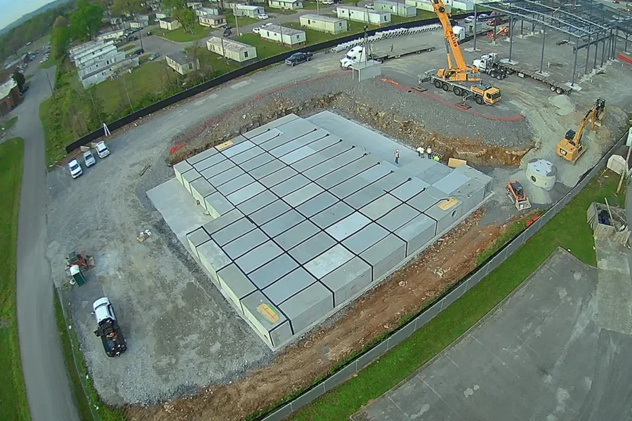 Aerial view of a construction site with a large concrete foundation, a crane, several vehicles, and nearby mobile homes.