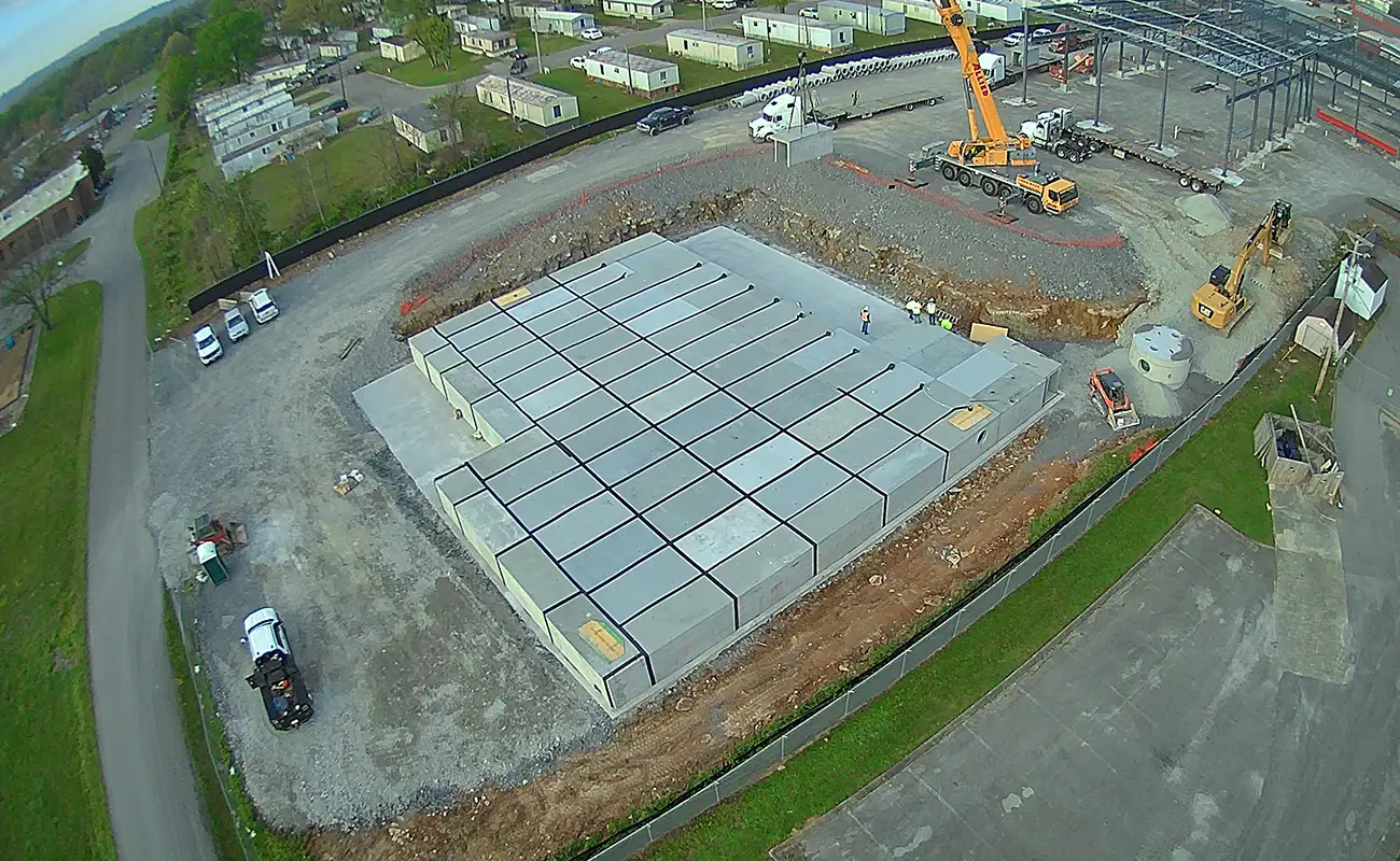 Aerial view of a construction site with a large concrete foundation, a crane, several vehicles, and nearby mobile homes.