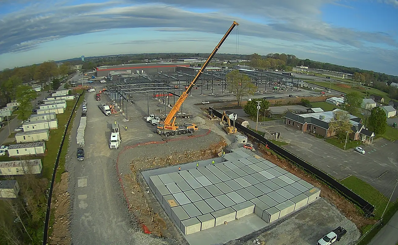 A large crane operates at a construction site with concrete slabs, surrounded by vehicles and temporary buildings, in a semi-urban area under a cloudy sky.