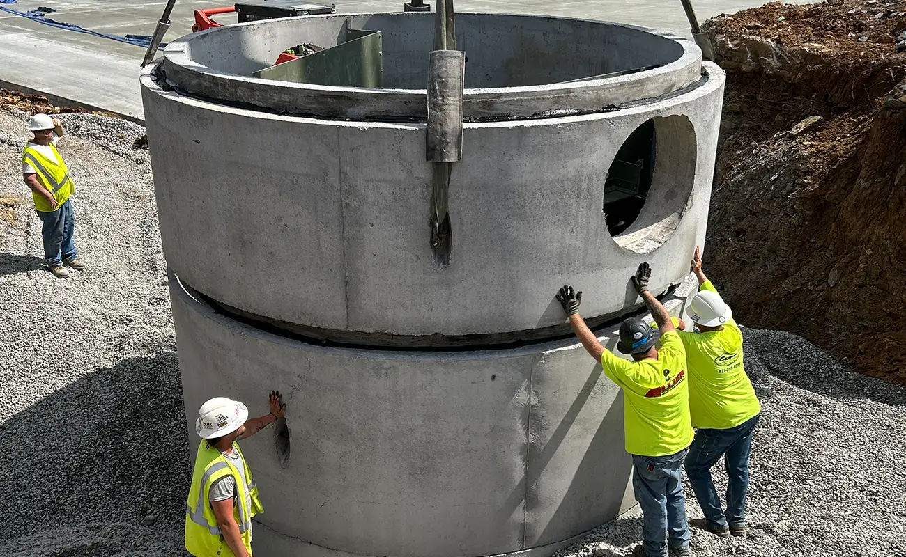 Three construction workers guide a large concrete cylinder section into place with a crane at a construction site, while a fourth worker observes.